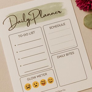 May include: A beige daily planner with the words "Daily Planner" in a handwritten font. The planner includes sections for a to-do list, schedule, daily bites, and a glow meter with emoji faces. The Grit & Glow Co. logo is at the bottom.