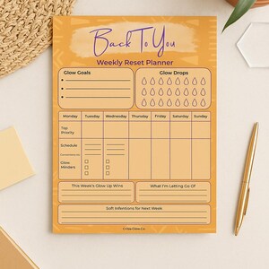 May include: An orange weekly reset planner with the text "Back To You" in purple script. The planner includes sections for glow goals, glow drops, and a weekly schedule. A gold pen rests on the white surface next to the planner.