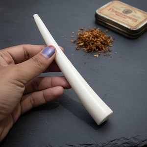 May include: A smooth, off-white smoking pipe with a tapered design. The pipe is held in a hand, with a small pile of tobacco and a vintage tin in the background. The pipe's length is approximately 6 inches.