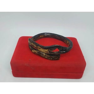 Könnte beinhalten: Ein dunkelbraunes und goldfarbenes Armband mit einem einzigartigen, organischen Design. Das Armband wird auf einer roten Samtbox präsentiert, die seine komplizierten Details und Kontrastfarben hervorhebt. Das Armband scheint aus Holz oder einem ähnlichen Material zu bestehen.