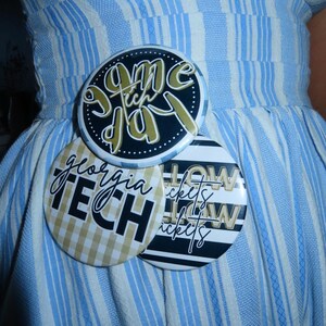 May include: Three round buttons with text and designs are pinned to a blue and white striped dress. The top button says "Game Day" in gold script on a black background. The middle button says "Georgia Tech" in black script on a gold and white checkered background. The bottom button says "How Tickets" in white script on a black and white striped background.