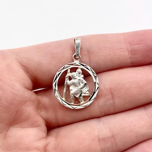 May include: A silver pendant featuring a detailed depiction of Saint Christopher carrying a child, within a geometric circular frame. The pendant has a small loop for a chain and is held in a hand against a white background.