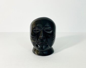 May include: A black, carved stone sculpture of a stylized human face. The face has defined features, including closed eyes, a prominent nose, and a detailed mouth. The sculpture is set against a plain white background.