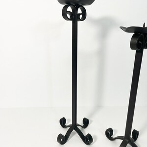 Pode incluir: Dois casti&ccedil;ais de metal preto com um design de rolagem. Os casti&ccedil;ais t&ecirc;m uma base alta e fina e uma parte superior larga e plana para segurar as velas.