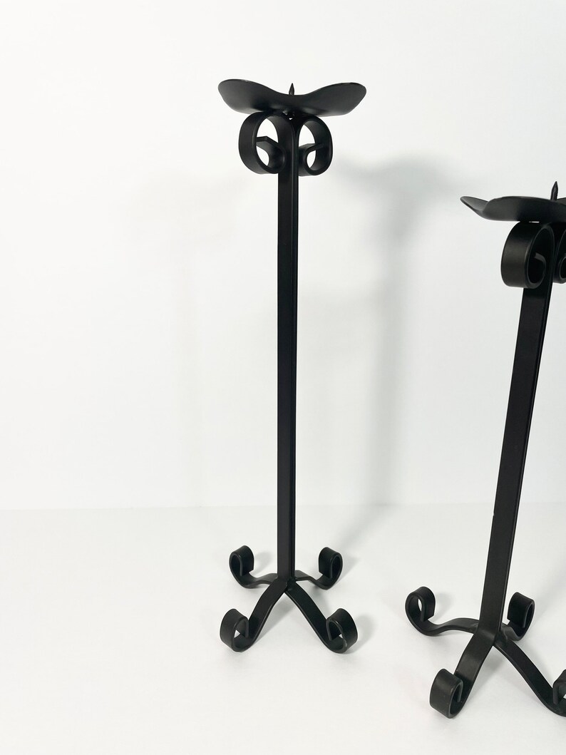 Pode incluir: Dois casti&ccedil;ais de metal preto com um design de rolagem. Os casti&ccedil;ais t&ecirc;m uma base alta e fina e uma parte superior larga e plana para segurar as velas.