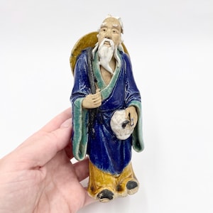 May include: A ceramic figurine of an elderly man with a long white beard, wearing a blue robe with green trim and a yellow hat. The figurine is holding a bag and a staff, and has a weathered appearance.