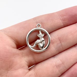 May include: A silver-colored charm featuring a seated elf figure within a circular frame. The elf has pointed ears and is holding its legs. The charm has a small loop at the top for attaching to a necklace or bracelet. The charm is likely made of metal.