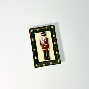 May include: A ceramic rectangular dish with a green border and gold stars. The center of the dish features a red, white, and black nutcracker holding a white mouse.