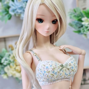 以下が含まれることがあります： 長い明るいブロンドの髪と青い目を持つ人形。人形は、薄緑色のリボンとレースのトリムが付いた花柄のブラジャーを着ています。人形はまた、長いぶら下がりのイヤリングをしています。
