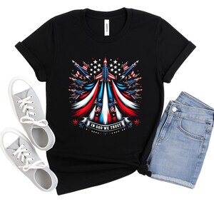 Peut inclure: T-shirt noir avec un motif patriotique de fusées, d'étoiles et de rayures rouges, blanches et bleues. Le texte "IN GOD WE TRUST" est affiché sur une bannière. Le t-shirt est associé à un short en jean et des baskets grises.