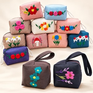 Puede incluir: Una colección de estuches rectangulares de tela en varios colores, incluyendo rosa, azul, marrón y blanco, cada uno con diseños bordados únicos de flores o setas. Algunos estuches tienen correas de muñeca y todos incluyen cierres de cremallera.