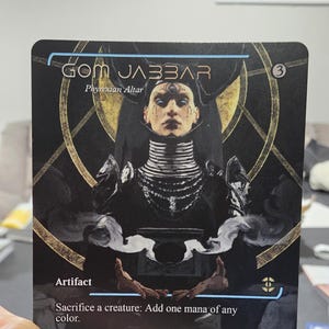 May include: A black and gold card with the text "GOM JABBAR" and "Phyrexian Altar." The card features an illustration of a person with a detailed neckpiece and the word "Artifact." The card also includes text from Gaius Helen Mohiam.