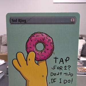 Op de afbeelding: Een aangepaste Sol Ring-kaart met een cartoonhand die een roze geglazuurde donut vasthoudt. De kaart heeft de tekst "TAP FOR 2? DON'T MIND IF I DO!" en de naam "Sol Ring" bovenaan.