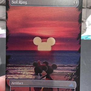 Può includere: Una carta Sol Ring con una scena di tramonto con sagome di Topolino e Minnie. La carta ha un bordo nero con la parola "Artifact" in basso. Il testo "Sol Ring" è in alto. Lo sfondo mostra un tramonto con una sagoma di Topolino.