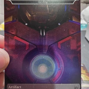 Può includere: Carta collezionabile "Arc Reactor" della serie Sol Ring. Presenta un design circolare centrale e la parola "Artifact" in basso. Lo sfondo è rosso scuro e viola con finitura metallica.