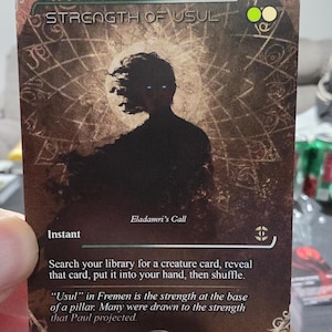 May include: A trading card with the title "STRENGTH OF USUL" and the text "Eladamri's Call". The card features a silhouetted figure with glowing eyes against a brown background with a circular design. The card's text describes the card's function.