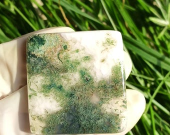 May include: A square, polished moss agate stone. The stone features a mix of white, green, and brown patterns, resembling a landscape. The agate is held in a gloved hand against a backdrop of green grass.