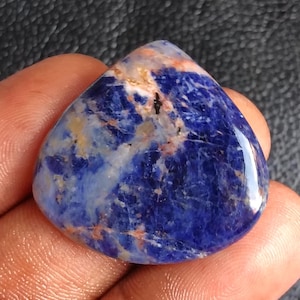 Peut inclure: Une pierre précieuse de sodalite polie, de forme triangulaire. La pierre présente une couleur de base bleu foncé avec des veines blanches, orange et marron. La surface est lisse et réfléchissante, mettant en valeur les motifs et couleurs naturels du minéral.