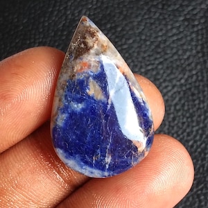 Puede incluir: Una gema en forma de lágrima con una superficie pulida. La piedra presenta una base azul oscuro con patrones arremolinados de blanco, marrón y naranja. La gema se sostiene en una mano, mostrando su belleza natural y variaciones de color únicas.