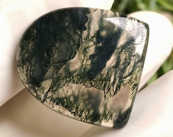 May include: A polished, semi-circular moss agate gemstone. The stone features a translucent base with intricate, tree-like patterns in shades of green and white. The gemstone is held in a gloved hand, showcasing its natural beauty.