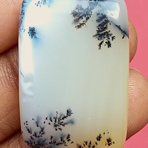 May include: A rectangular, polished gemstone with a creamy white base and intricate black and blue dendritic inclusions. The stone has rounded edges and a smooth, glossy surface. The overall effect is reminiscent of a miniature landscape.