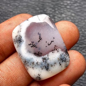 May include: A polished, rectangular dendritic agate gemstone featuring a smooth, translucent white and pale lavender surface. The stone displays intricate, dark black, fern-like branching inclusions that spread across the centre and edges.