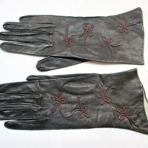 May include: A pair of dark brown leather gloves featuring decorative stitching in a deep red thread. The gloves are adorned with embroidered bow-like motifs on the back of the hand, showcasing a smooth finish and a classic five-finger design.