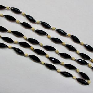May include: A long necklace featuring alternating black and cream-colored beads. The black beads are faceted, giving them a sparkling appearance. The necklace is laid out on a white surface, showcasing its length and design.