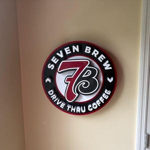 Puede incluir: Un letrero redondo con las palabras "SEVEN BREW DRIVE THRU COFFEE" en letras blancas. El letrero tiene un borde rojo y negro, con un centro blanco con el número "7" y la letra "B".