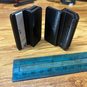 May include: Two black 3D-printed plastic clips with a Z-shaped profile, each featuring a white adhesive strip on one side. A blue plastic ruler is positioned in front, showing the clips are approximately 1.5 inches wide at their base.