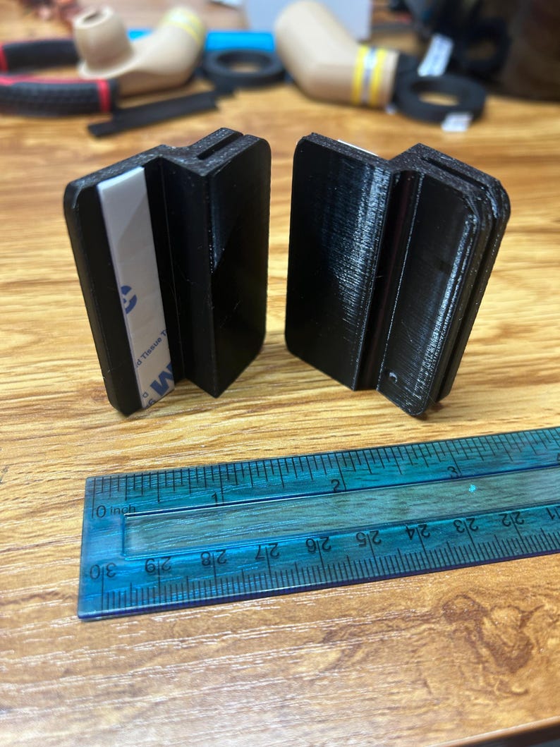 May include: Two black 3D-printed plastic clips with a Z-shaped profile, each featuring a white adhesive strip on one side. A blue plastic ruler is positioned in front, showing the clips are approximately 1.5 inches wide at their base.