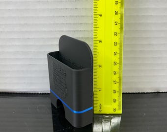 May include: A black 3D-printed phone holder with a blue accent line. The holder is approximately 3 inches tall and features a design on the side. A yellow ruler is placed next to the holder for scale.