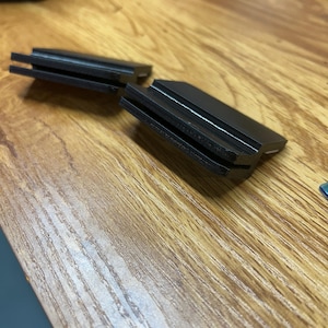 May include: Two black, rectangular plastic clips featuring a horizontal slot running through the center of each piece. These components have a smooth, matte finish and a stepped design, with one side thicker than the other, creating a distinct profile.