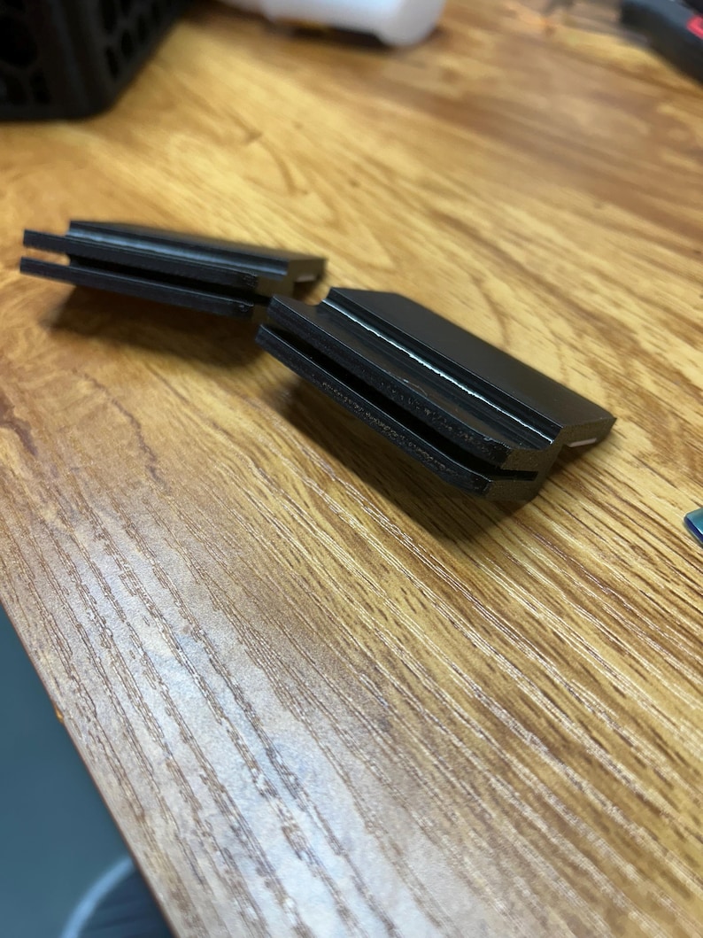 May include: Two black, rectangular plastic clips featuring a horizontal slot running through the center of each piece. These components have a smooth, matte finish and a stepped design, with one side thicker than the other, creating a distinct profile.