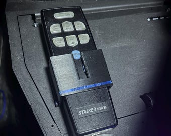 May include: A black remote control with white buttons is held in a 3D-printed holder. The remote has the text "STALKER DSR 2X" and "JOTTO DESK" is visible. The holder is attached to a black surface with USB ports.