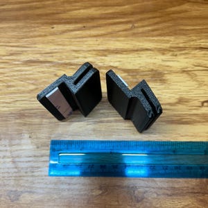 May include: Two black, L-shaped plastic mounting brackets with textured surfaces and white adhesive strips on the inner faces. A blue plastic ruler is positioned below the brackets, showing they measure approximately 1.5 inches in width.