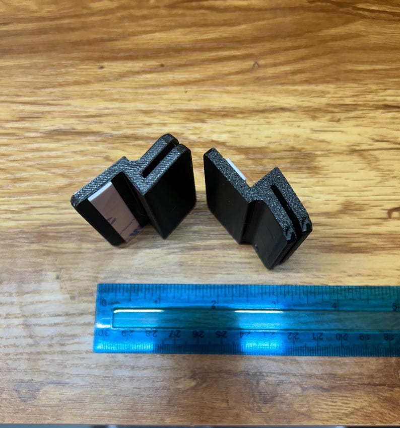 May include: Two black, L-shaped plastic mounting brackets with textured surfaces and white adhesive strips on the inner faces. A blue plastic ruler is positioned below the brackets, showing they measure approximately 1.5 inches in width.