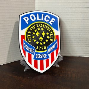 May include: A shield-shaped Police emblem featuring the words "POLICE" and "CITY OF LOUISVILLE" in blue and yellow. The emblem includes the words "COURAGE", "SERVICE", and "INTEGRITY" in red and white, with the year "1778".