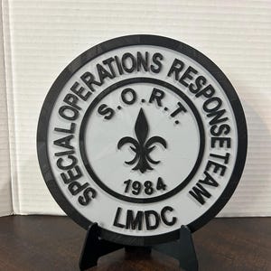 May include: A round, black and white emblem with the text "SPECIAL OPERATIONS RESPONSE TEAM" encircling the edge. The center features a fleur-de-lis and the text "S.O.R.T. 1984 LMDC". The emblem is displayed on a black stand.
