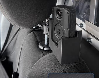 May include: A black Axon body camera transport mount attached to a vehicle headrest. The text reads: NEW PRODUCT AXON BODY CAMERA TRANSPORT MOUNT, KEEP THE REAR SEAT ON CAMERA, Positions your Axon body camera to record people in custody during transport.