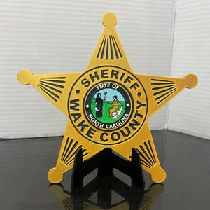 May include: A gold-colored, five-pointed star-shaped badge with the words "SHERIFF STATE OF WAKE COUNTY NORTH CAROLINA" in black lettering. The center of the badge features a circular emblem with a depiction of two figures and a landscape. The badge is displayed on a black stand.