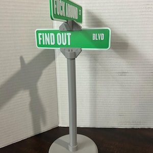 以下が含まれることがあります： 「FUCK AROUND ST」と「FIND OUT BLVD」と書かれた緑色の標識が付いた、斬新な街路標識。標識は灰色のポールとベースに取り付けられています。この標識はユーモラスな装飾品です。