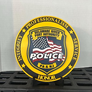 Op de afbeelding: Een rond embleem van de Delaware River Port Authority Police met een gele rand en de woorden Integrity, Professionalism, Service en Honor. Het centrale schild toont een Amerikaanse vlag, de tekst Police en PA & NJ in het geel.