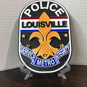 May include: A black and white Louisville Metro Police badge with the words "POLICE" and "LOUISVILLE" in white lettering. The badge features a gold fleur-de-lis and the words "SERVICE METRO INTEGRITY" in blue.