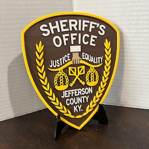 May include: A brown and yellow Sheriff's Office sign with the text "SHERIFF'S OFFICE" at the top. The sign also includes the words "JUSTICE" and "EQUALITY" and "JEFFERSON COUNTY KY."