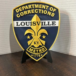 May include: A shield-shaped emblem for the Louisville Department of Corrections. The emblem features gold lettering and accents on a navy blue background. The text includes "DEPARTMENT OF CORRECTIONS", "LOUISVILLE", "KY METRO", and "1778".