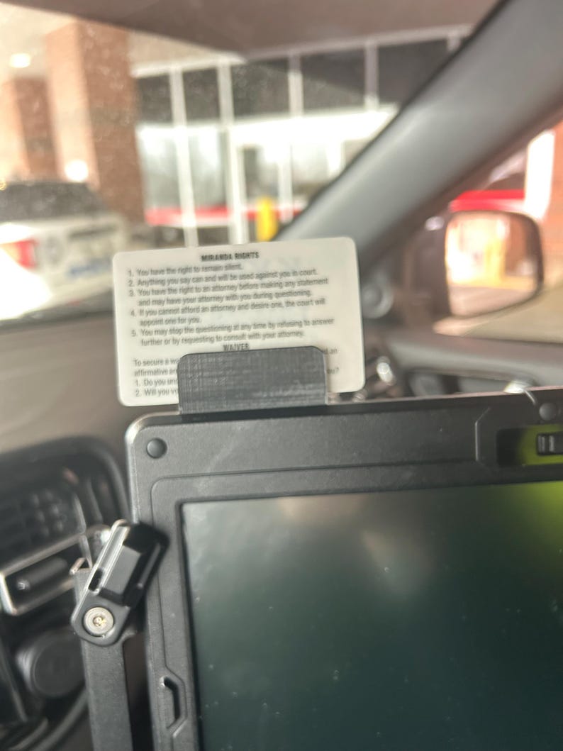 May include: A white rectangular card featuring the printed text Miranda Rights and a waiver, held in place by a black plastic clip mounted on the top edge of a black electronic device screen. The card lists five numbered legal rights.
