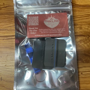 May include: A silver metallic resealable pouch containing a dark gray rectangular object with a thin blue line and a blue metal bottle opener on a keyring. A red card is inside with the text Thin Line 3D Printing & Laser Engraving.
