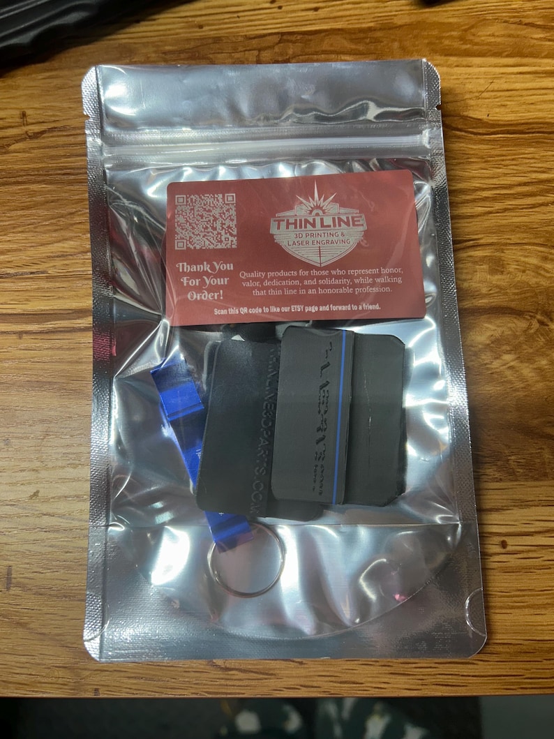 May include: A silver metallic resealable pouch containing a dark gray rectangular object with a thin blue line and a blue metal bottle opener on a keyring. A red card is inside with the text Thin Line 3D Printing & Laser Engraving.