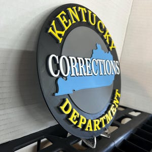 May include: A circular black sign featuring a blue silhouette of the state of Kentucky in the center. The text KENTUCKY is arched in yellow at the top, CORRECTIONS is in white across the center, and DEPARTMENT is arched in yellow at the bottom.